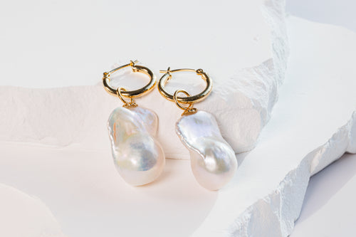 A pair of baroque pearl earrings with gold hoops, displayed on a white surface with a subtle background.